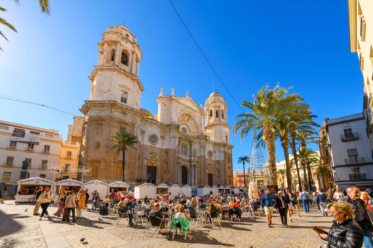 Place animée à Cadix avec la cathédrale, de nombreuses personnes assises aux terrasses, palmiers et architecture historique sous un ciel bleu
