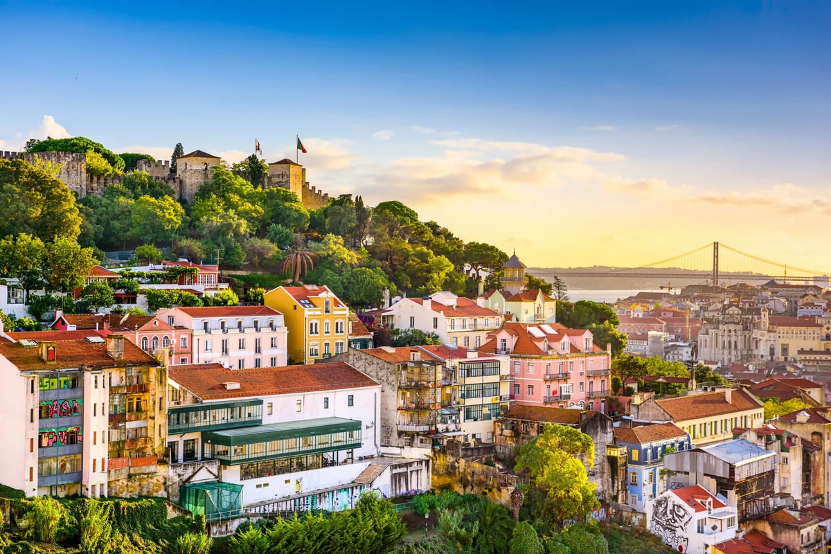Blick über die Altstadt von Lissabon mit bunten Häusern, Burg auf dem Hügel und Hängebrücke im Hintergrund bei Abendlicht