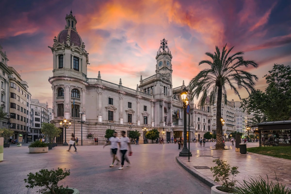 Bâtiment historique à Valence au crépuscule avec des lampadaires allumés, des palmiers et des passants sur une grande place
