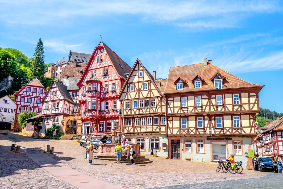 Place ensoleillée à Miltenberg avec maisons à colombages colorées, fontaine et passants sur des pavés devant des façades historiques.