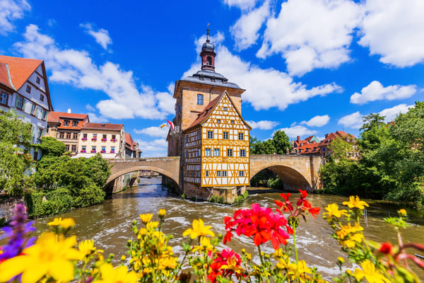 Altes Rathaus in Bamberg auf einer Brücke über dem Fluss, eingerahmt von Blumen im Vordergrund und Altstadt unter blauem Himmel.