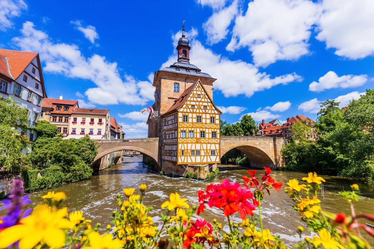 Ancien hôtel de ville de Bamberg posé sur un pont au-dessus de la rivière, encadré par des fleurs au premier plan et la vieille ville sous un ciel bleu.