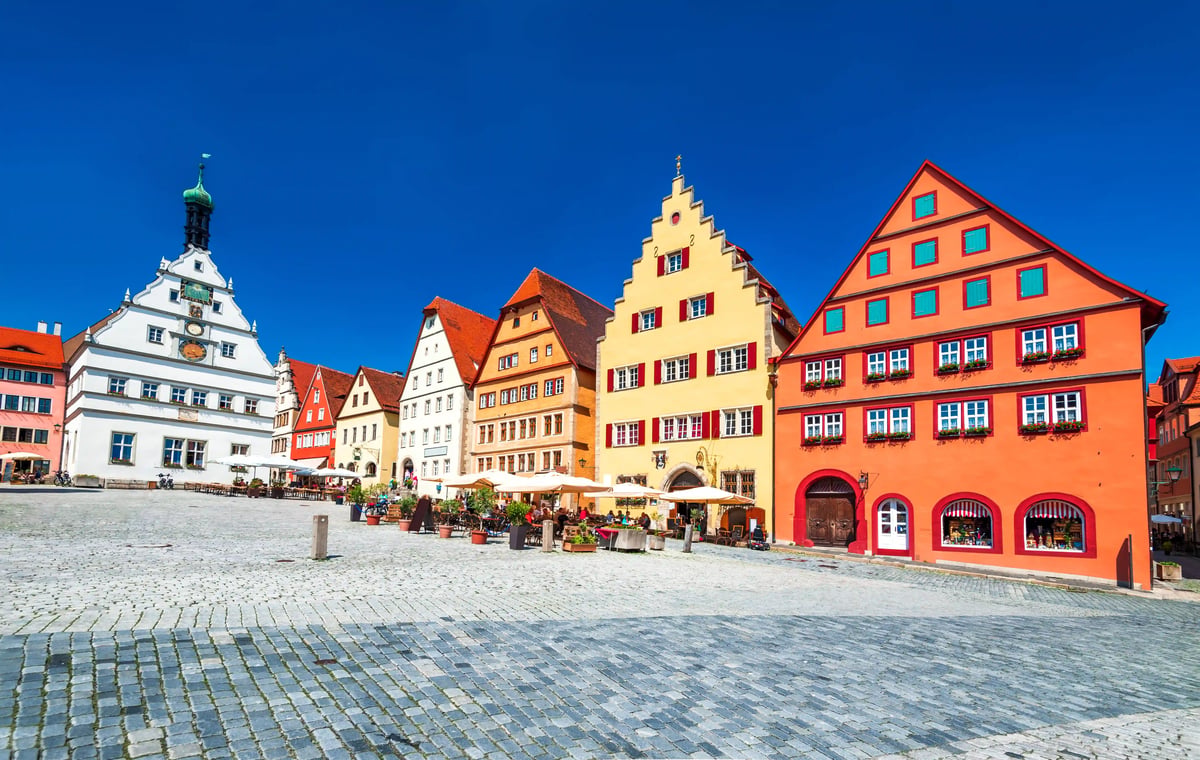 Place de marché colorée à Rothenburg ob der Tauber avec des maisons à pignons historiques, des terrasses et des passants sur les pavés, sous un ciel bleu intense.