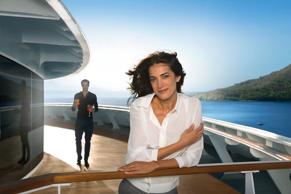 Frau steht entspannt an der Reling eines modernen Kreuzfahrtschiffes mit Blick auf das Meer, im Hintergrund eine weitere Person und eine ruhige Küstenlandschaft