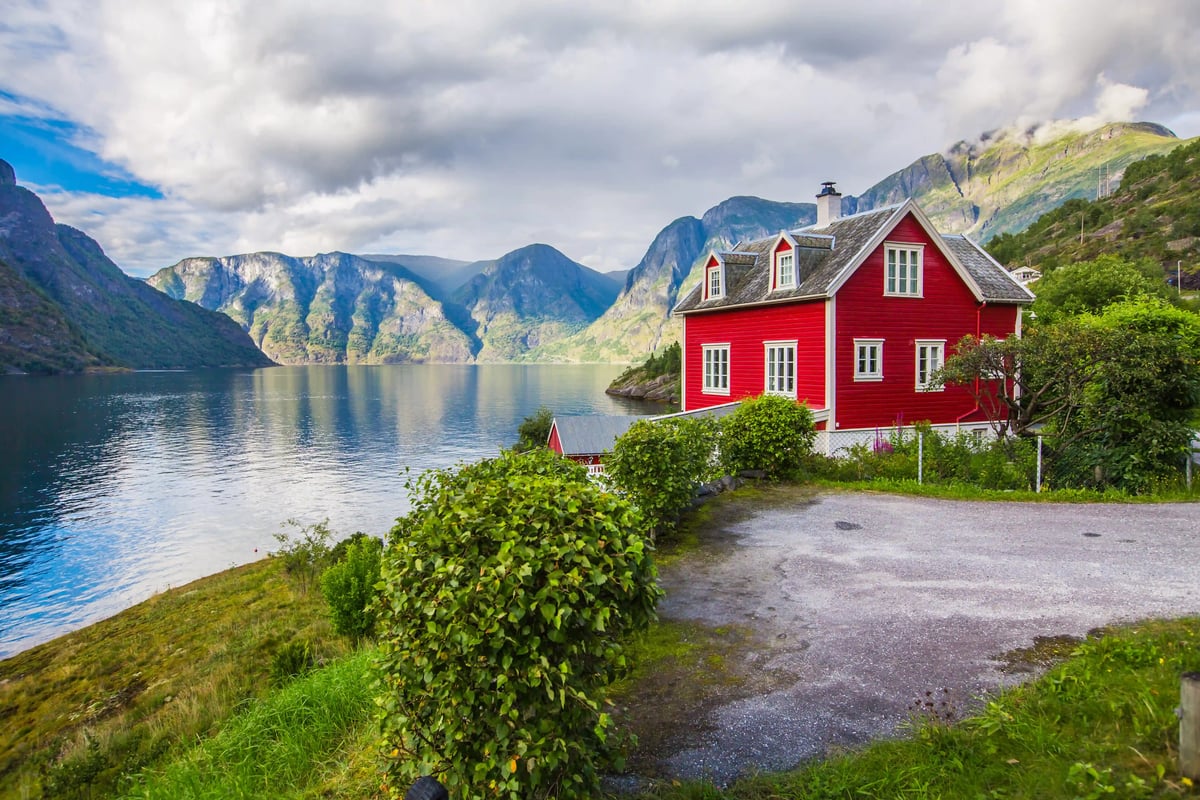 Rotes Holzhaus mit Garten steht am Ufer eines stillen Fjords, im Hintergrund steile Berge und grün bewachsene Hänge