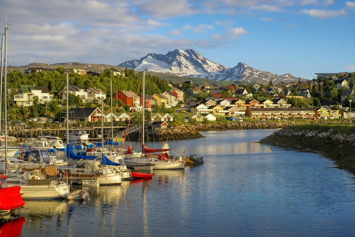 Ein idyllischer Bootshafen in Narvik mit Segelbooten im Vordergrund, bunten Holzhäusern am Hang und einem schneebedeckten Bergmassiv unter blauem Himmel