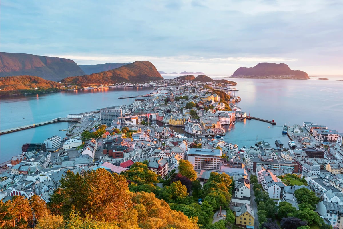 Luftaufnahme der norwegischen Stadt Ålesund bei Abenddämmerung, die sich auf einer schmalen Landzunge im blauen Meer erstreckt, umgeben von Bergen
