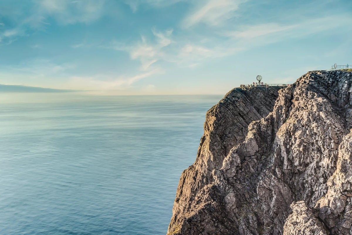 Die steile, felsige Klippe des Nordkaps in Norwegen über dem weiten Meer unter leicht bewölktem blauem Himmel mit der markanten Globus-Skulptur auf dem Plateau