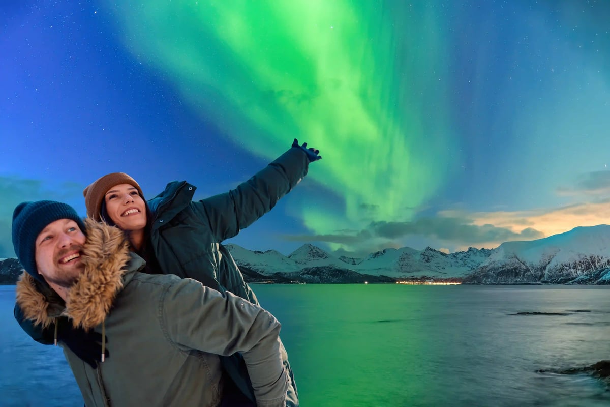Ein glückliches Paar in Winterkleidung vor einer nächtlichen Fjordlandschaft mit Bergen, während am Himmel leuchtend grüne Polarlichter tanzen