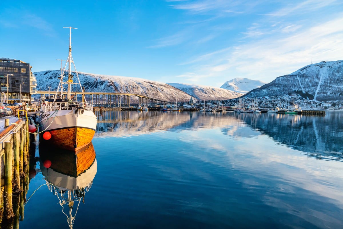 Ein hölzernes Fischerboot liegt im ruhigen Hafen von Tromsø, im Hintergrund spiegeln sich die Brücke und verschneite Berge im klaren, blauen Wasser