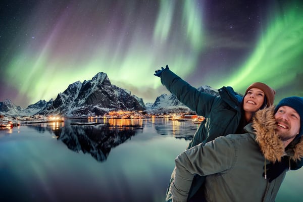 Polarlichter leuchten über einem Küstendorf mit Bergen, Häusern und Spiegelungen im Wasser bei Nacht
