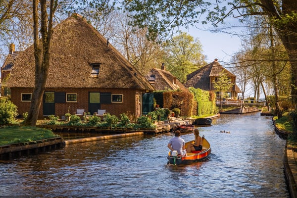 Boot mit zwei Personen fährt durch einen Kanal in Giethoorn vorbei an Reetdachhäusern und Bäumen.