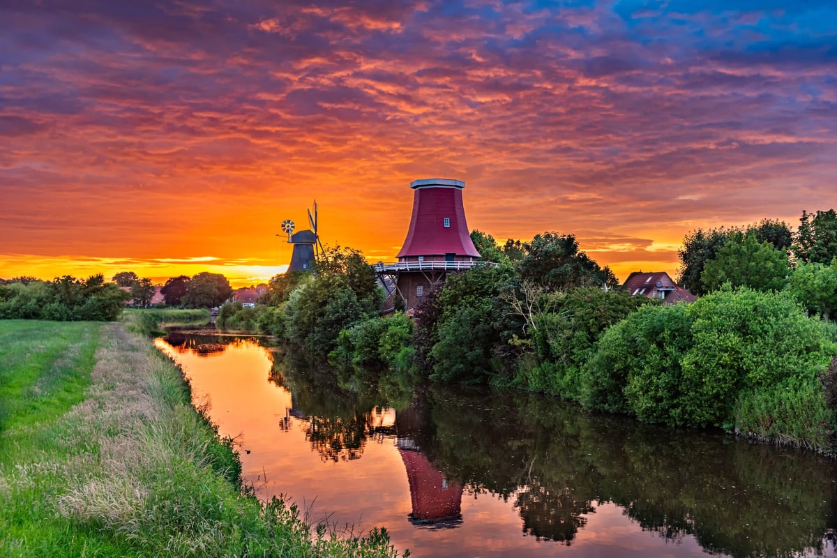 Zwillingsmuehlen von Greetsiel spiegeln sich bei Sonnenuntergang in einem ruhigen Wasserlauf.