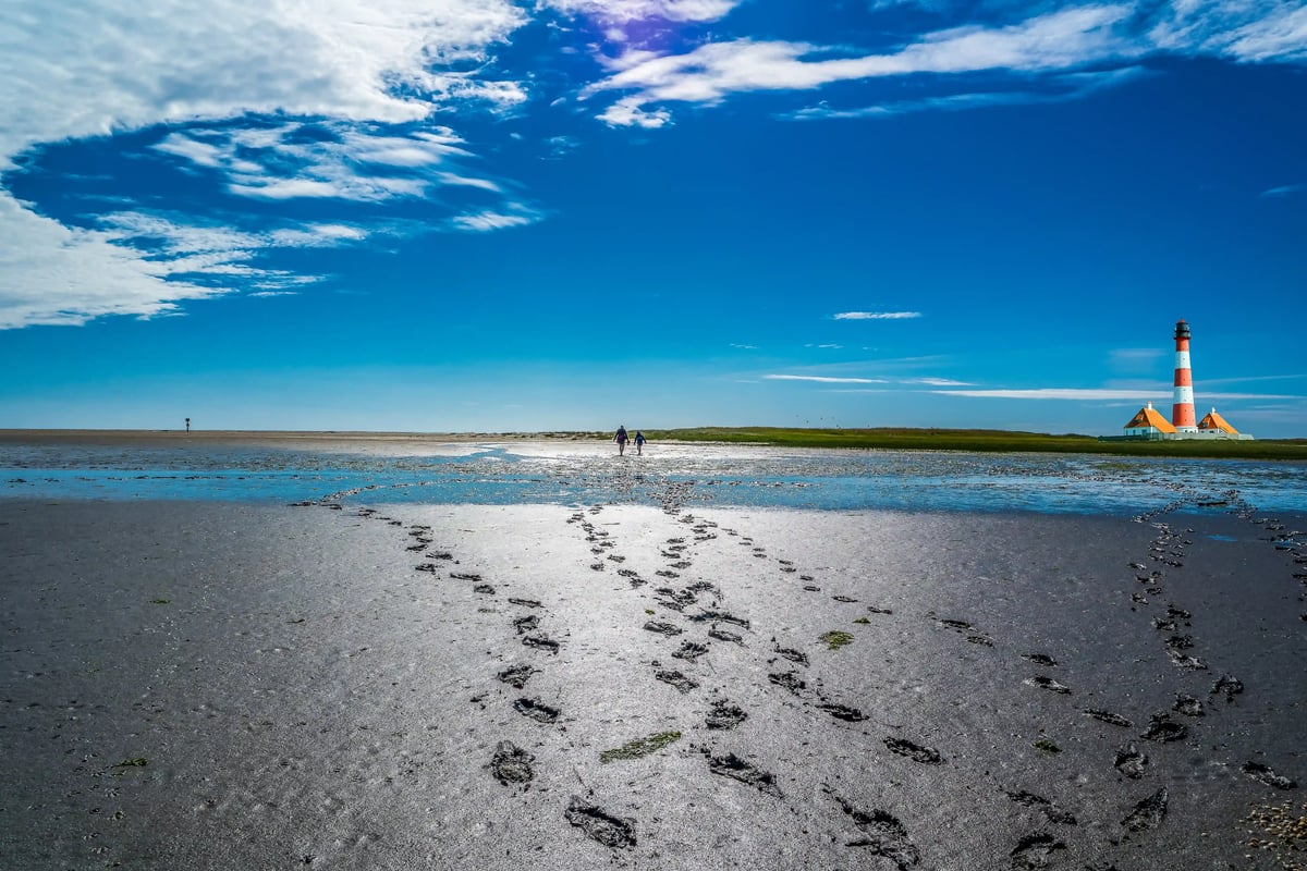 Weite Wattlandschaft mit Fußspuren im Schlick und Leuchtturm am Horizont