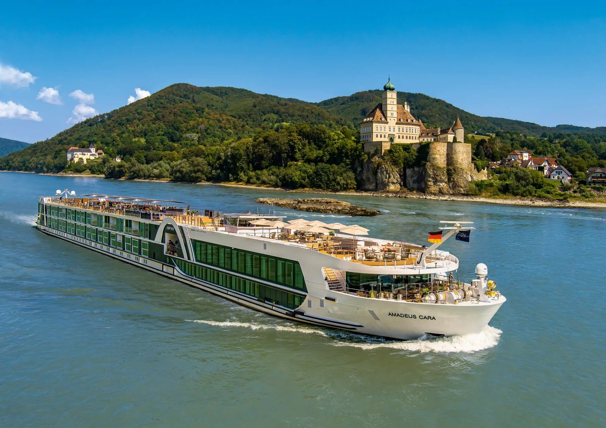 Flusskreuzfahrtschiff MS Amadeus Cara auf einem breiten Fluss vor einer Burganlage und bewaldeten Hügeln.