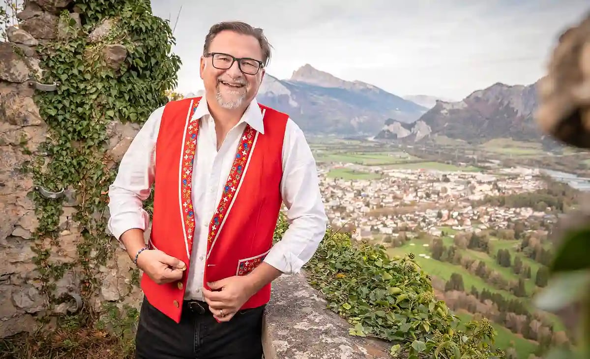 Mann in roter Weste steht auf einer Aussichtsterrasse vor einer Berglandschaft und blickt lächelnd in die Kamera.