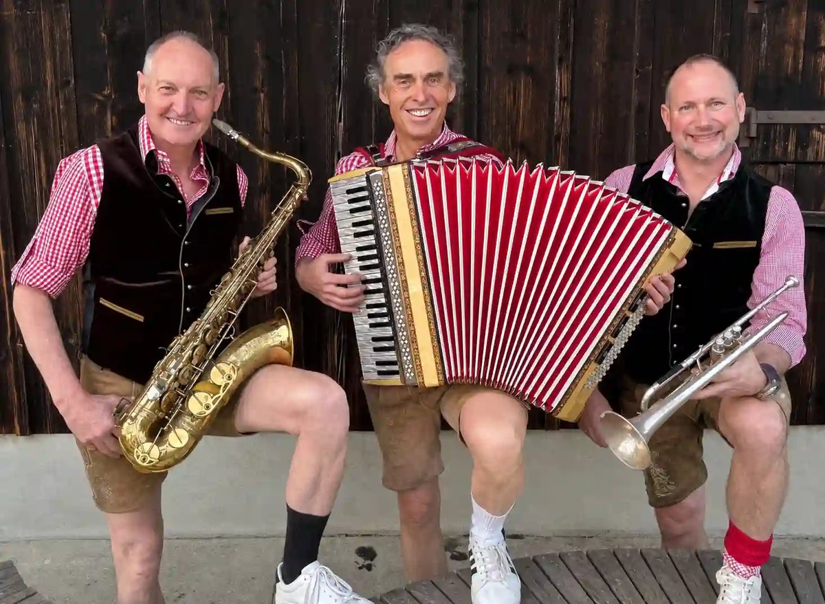 Drei Musiker in traditioneller Kleidung mit Saxofon, Akkordeon und Trompete vor einer Holzwand.