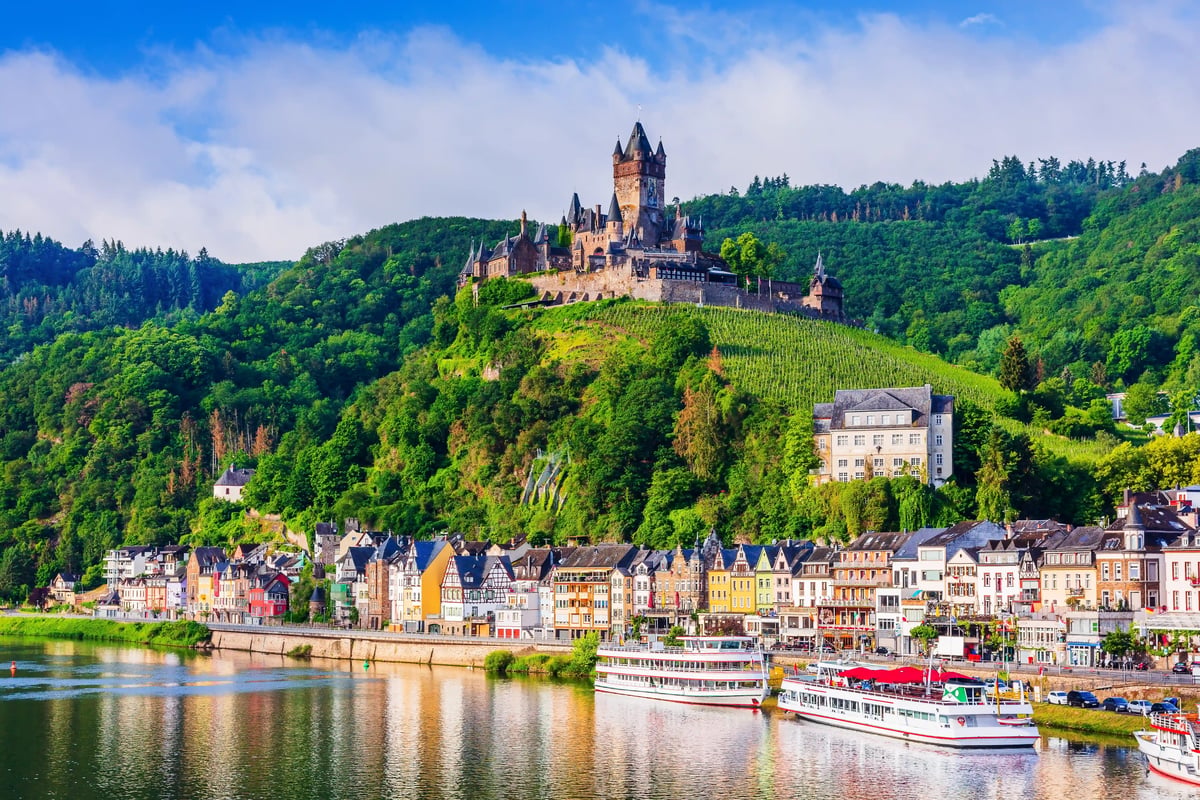 Blick auf Cochem an der Mosel mit Reichsburg auf einem bewaldeten Hügel und Schiffen am Flussufer.