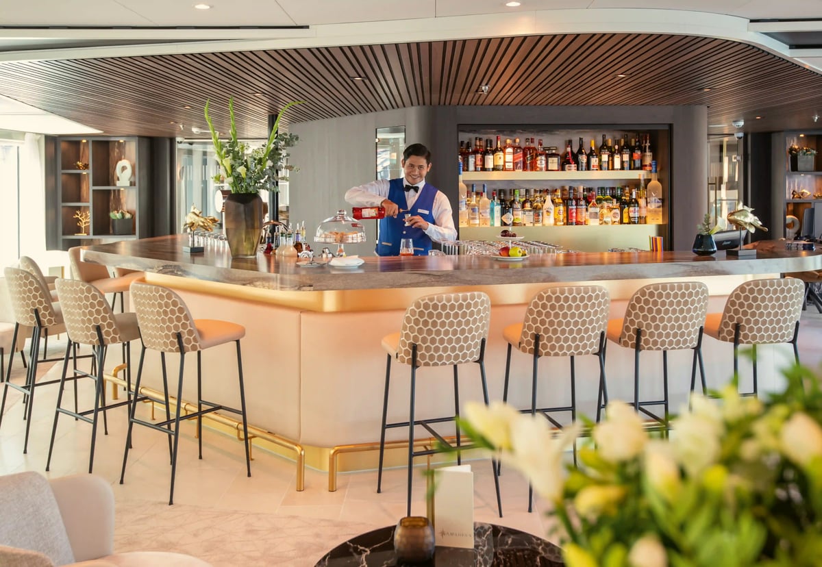 Elegante Bar auf dem Flusskreuzfahrtschiff MS AMADEUS Cara mit Sitzplätzen, beleuchteter Getränkeauswahl und Barkeeper bei der Zubereitung eines Getränks.