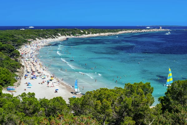 Beliebter Strand auf Ibiza mit Badegästen, türkisblauem Wasser und grüner Küstenlandschaft