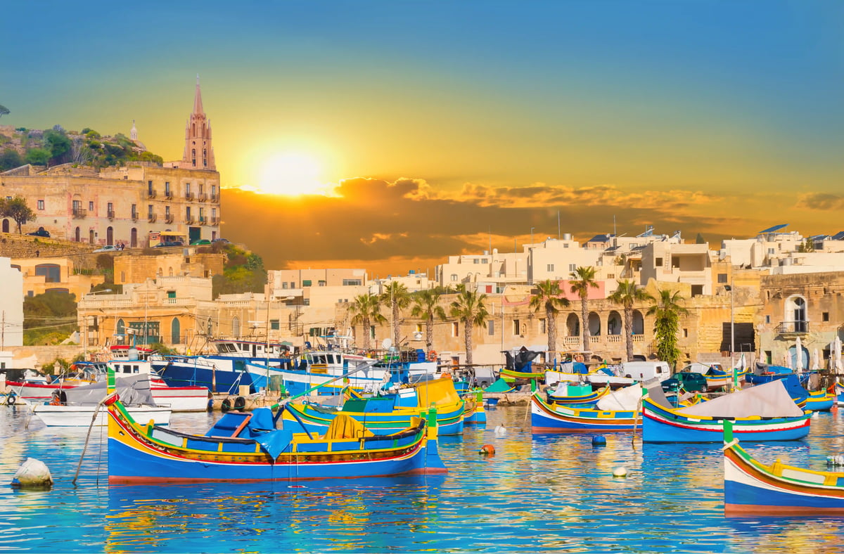 Bunte Fischerboote liegen im Hafen von La Valletta bei Sonnenuntergang, historische Gebäude im Hintergrund