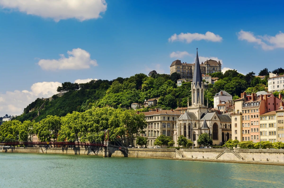 Vue des quais de Lyon avec église, bâtiments historiques et colline verdoyante.