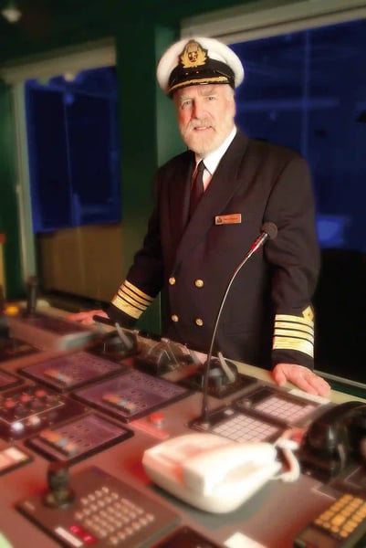 Kapitän in Uniform steht auf der Schiffsbrücke vor Steuerpulten und blickt freundlich in die Kamera