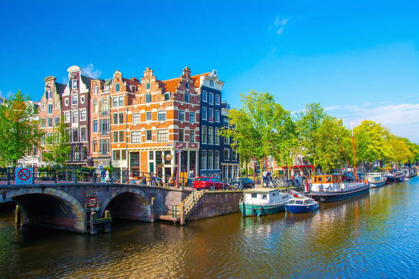 Amsterdamer Gracht mit Steinbrücke, schmalen Häusern und Booten am Ufer, sonniger Himmel und lebendige Strassenszene