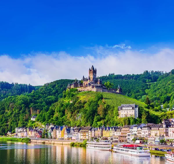 Burg auf einem grünen Hügel über der Mosel, darunter bunte Häuser am Flussufer und Ausflugsschiffe auf dem Wasser.