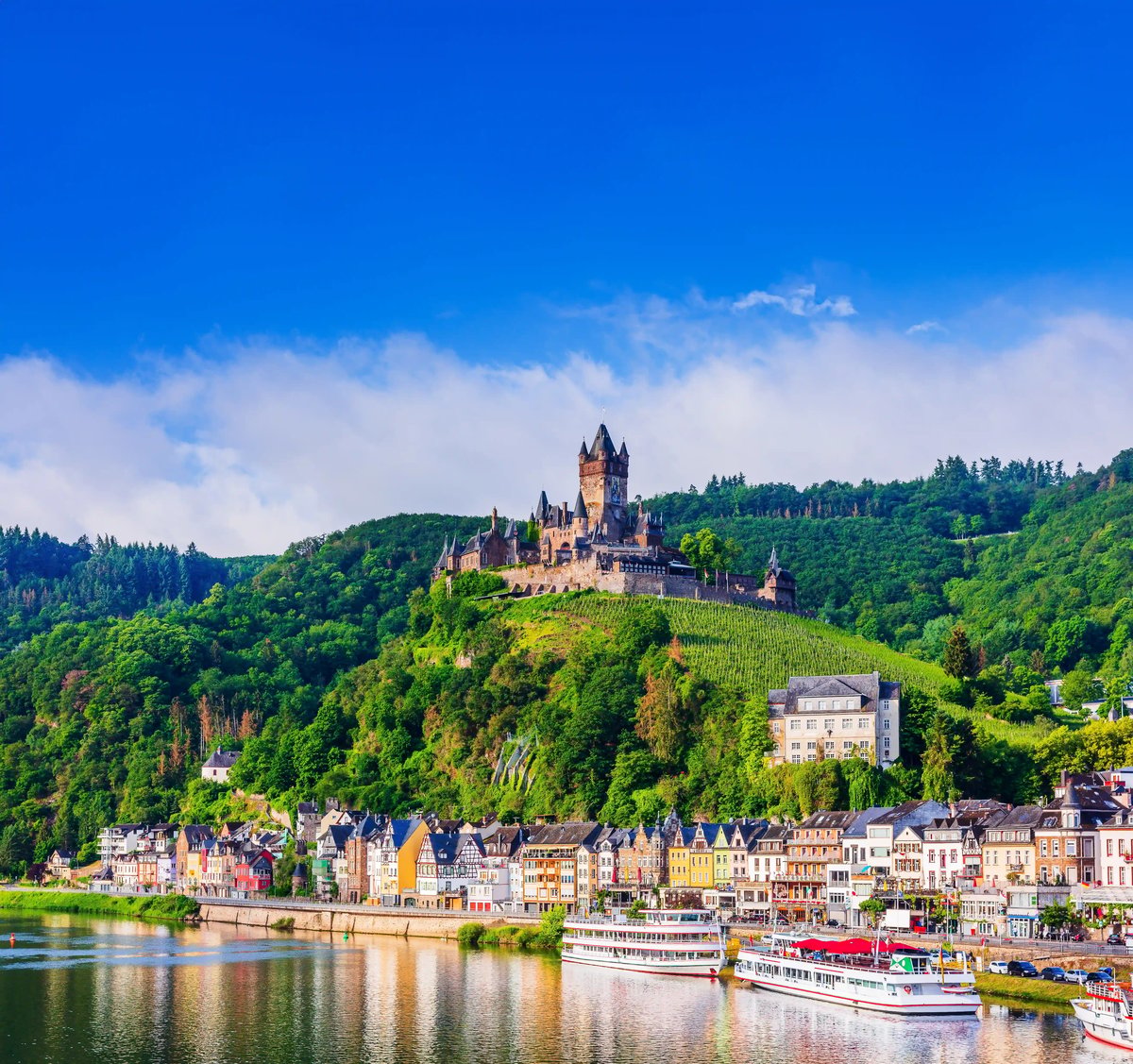 Burg auf einem grünen Hügel über der Mosel, darunter bunte Häuser am Flussufer und Ausflugsschiffe auf dem Wasser.