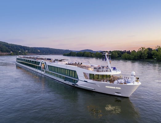 Modernes Flusskreuzfahrtschiff Amadeus Riva fährt bei Sonnenuntergang auf breitem Fluss, sanfte Hügel und Bäume am Ufer.
