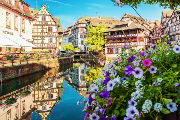 Fachwerkhäuser und Restaurants am Kanal in Strassburg, bunte Blumen im Vordergrund und Spiegelungen im Wasser.