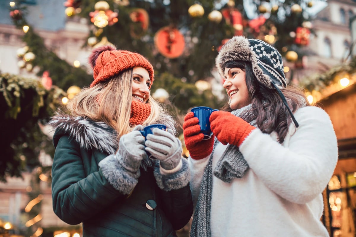 Deux femmes en vetements d hiver se tiennent sur un marche de Noel et sourient en tenant des tasses de boisson chaude devant un sapin illumine