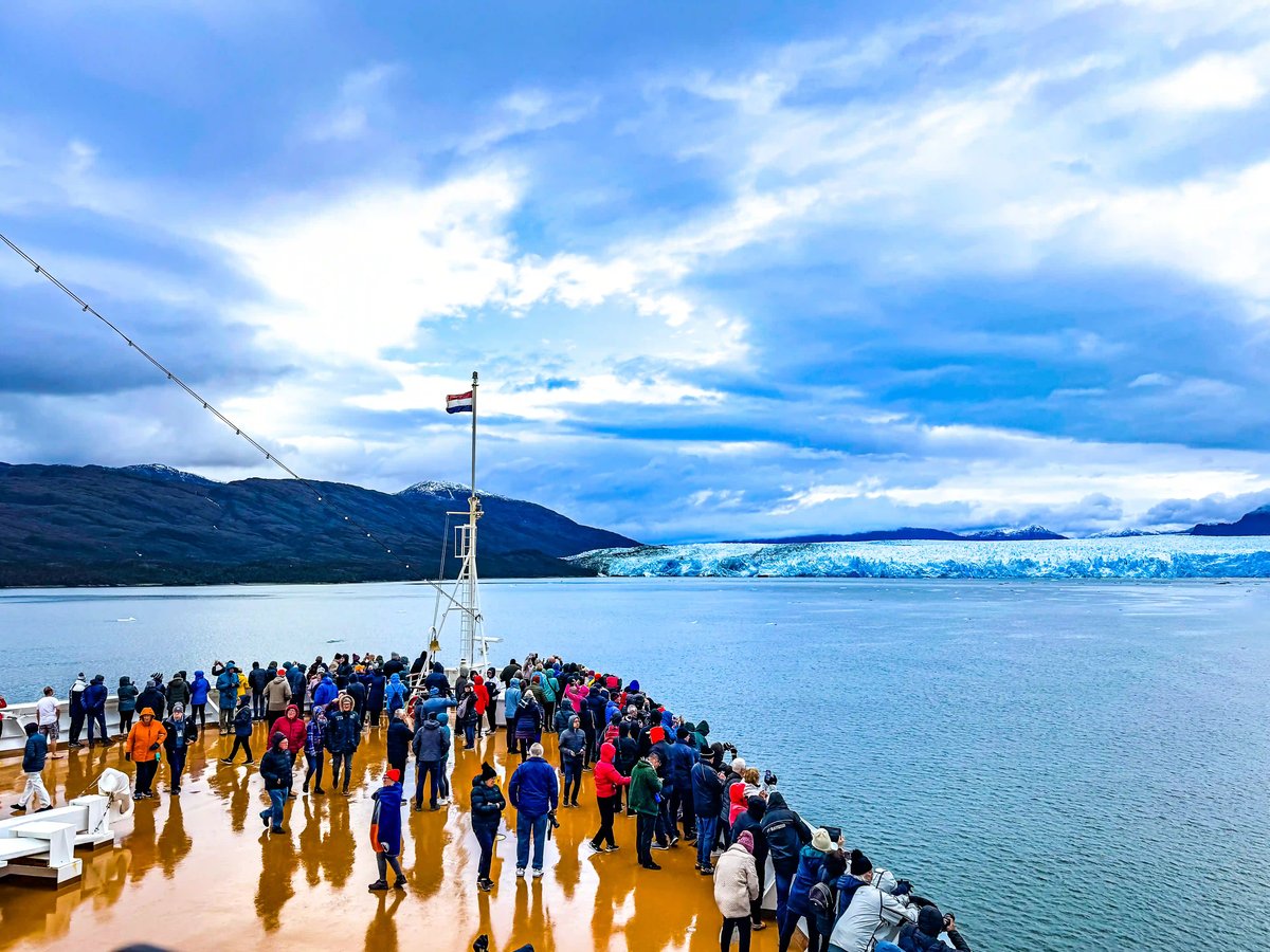 Viele Passagiere auf dem Aussichtsdeck eines Schiffes blicken auf eine breite, blau leuchtende Gletscherfront im Meer