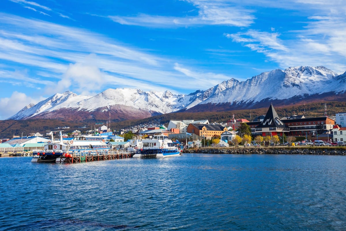 Hafen von Ushuaia mit Booten an einem Steg vor bunten Gebäuden und schneebedeckten Bergen unter blauem Himmel.
