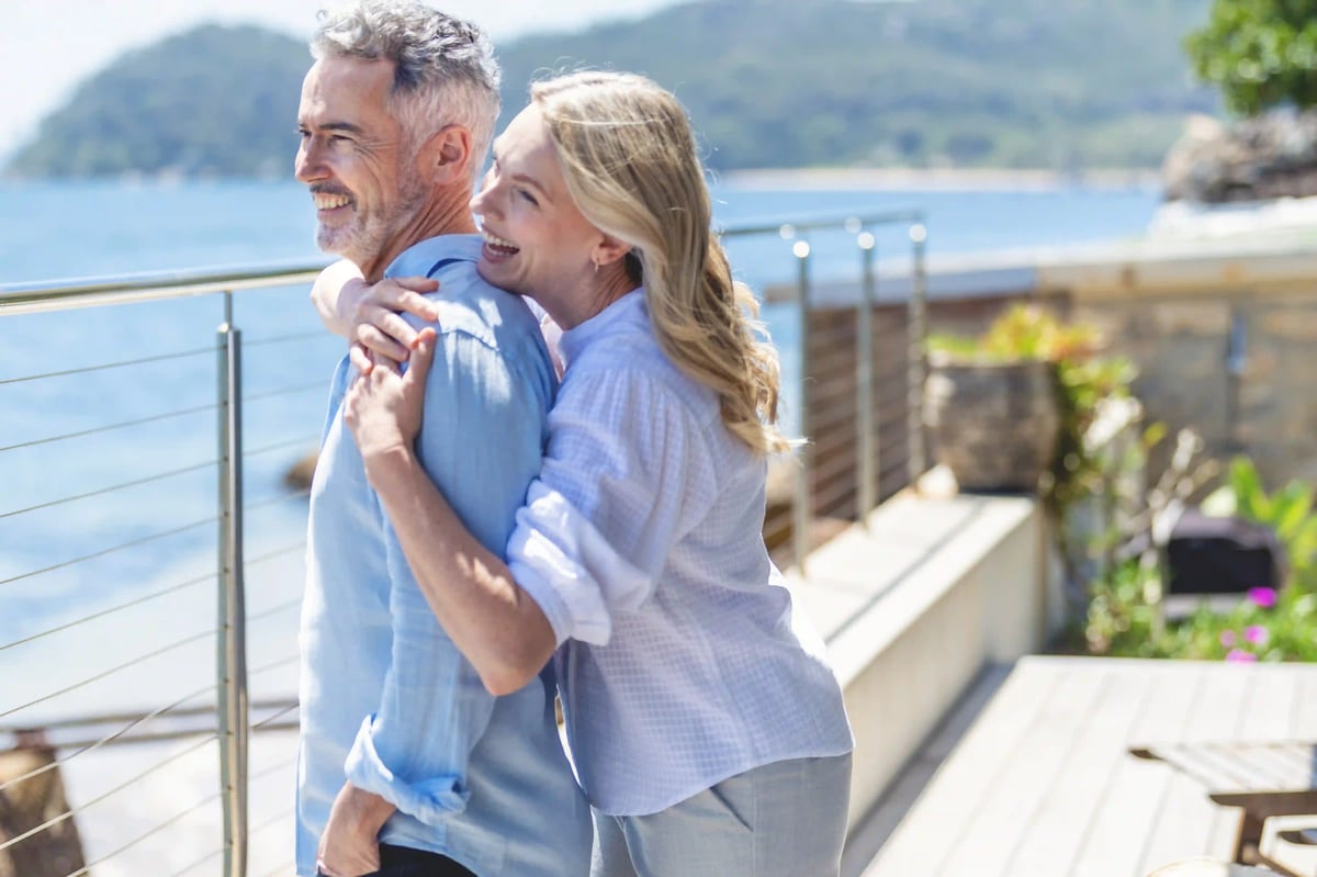 Lachendes Paar steht umarmt auf einer Terrasse mit Blick auf das Meer und eine grüne Küstenlandschaft