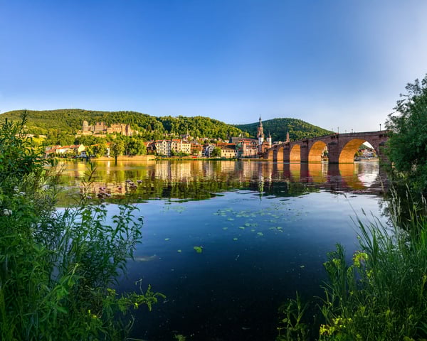 Blick über den Neckar auf die Alte Brücke in Heidelberg, mit Altstadt und bewaldeten Hügeln im warmen Abendlicht