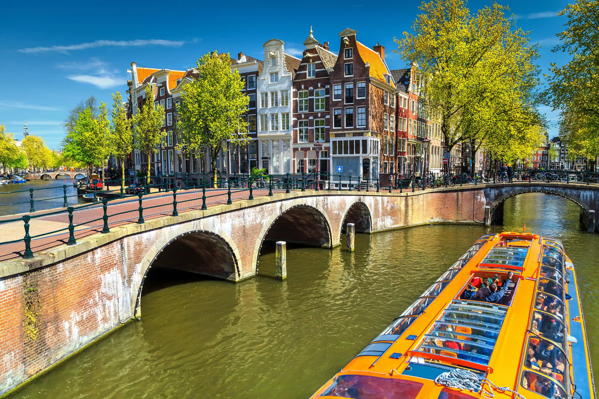Grachtenboot fährt unter einer gemauerten Bogenbrücke in Amsterdam, flankiert von hohen Giebelhäusern und Bäumen