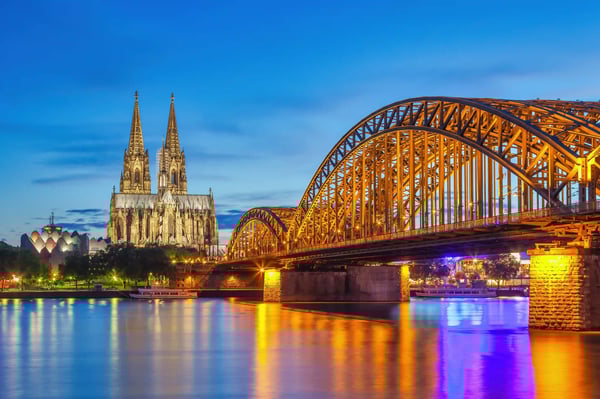 Kölner Dom neben einer grossen Stahlbogenbrücke am Abend, Lichter spiegeln sich im Rhein
