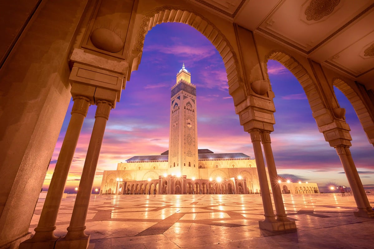 Beleuchtete Hassan-II.-Moschee in Casablanca bei Sonnenuntergang, gesehen durch steinerne Bögen