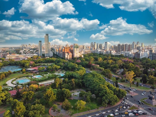 Blick über einen grossen Stadtpark mit vielen Bäumen auf eine dichte Skyline aus Hochhaeusern unter blauem Himmel mit Wolken