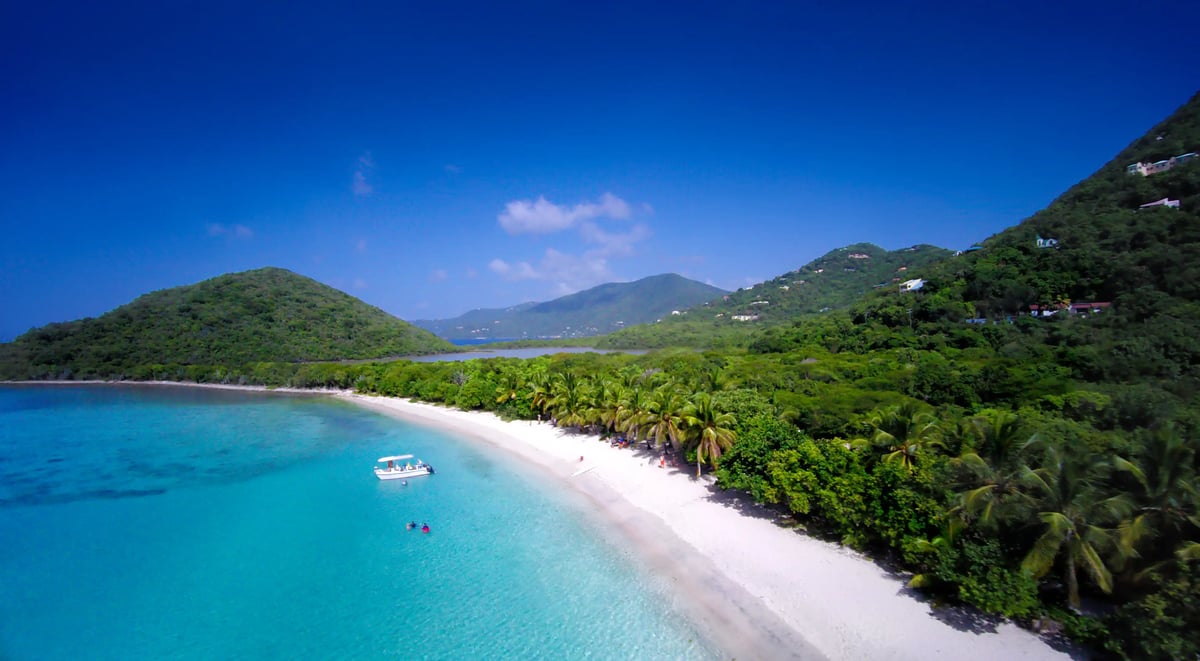 Luftaufnahme eines karibischen Strandes mit hellem Sand, türkisfarbenem Wasser, Palmen entlang der Küste und bewaldeten Hügeln im Hintergrund