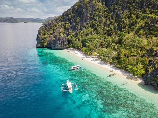 Tropischer Strand mit türkisfarbenem Wasser, Palmen, Kalksteinfelsen und kleinen Booten