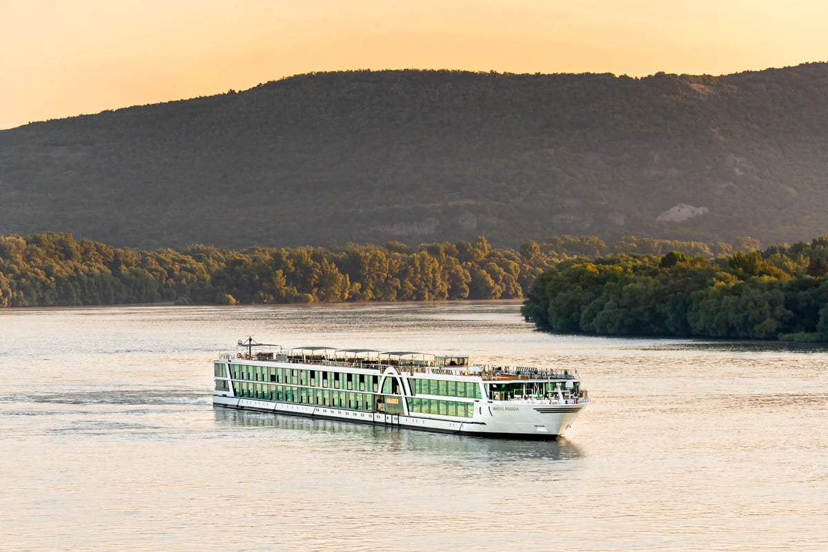 Navire fluvial moderne naviguant sur un fleuve calme devant des collines boisées au coucher du soleil