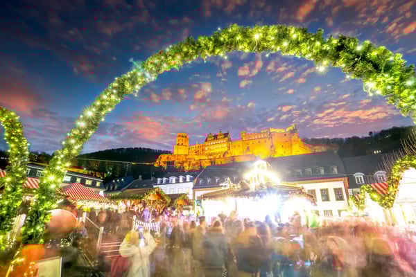 Heidelberger Schloss über dem Weihnachtsmarkt bei Schneefall, Lichterbogen und Besucher in winterlicher Abendstimmung.