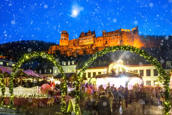 Heidelberger Schloss über dem Weihnachtsmarkt bei Schneefall, Lichterbogen und Besucher in winterlicher Abendstimmung.