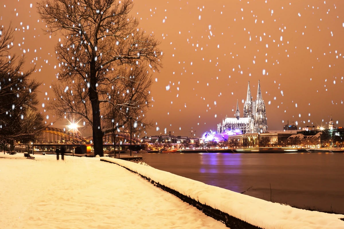 Verschneiter Rheinuferspazierweg in Köln bei Nacht, Dom und Brücke im Hintergrund, Schneeflocken fallen.