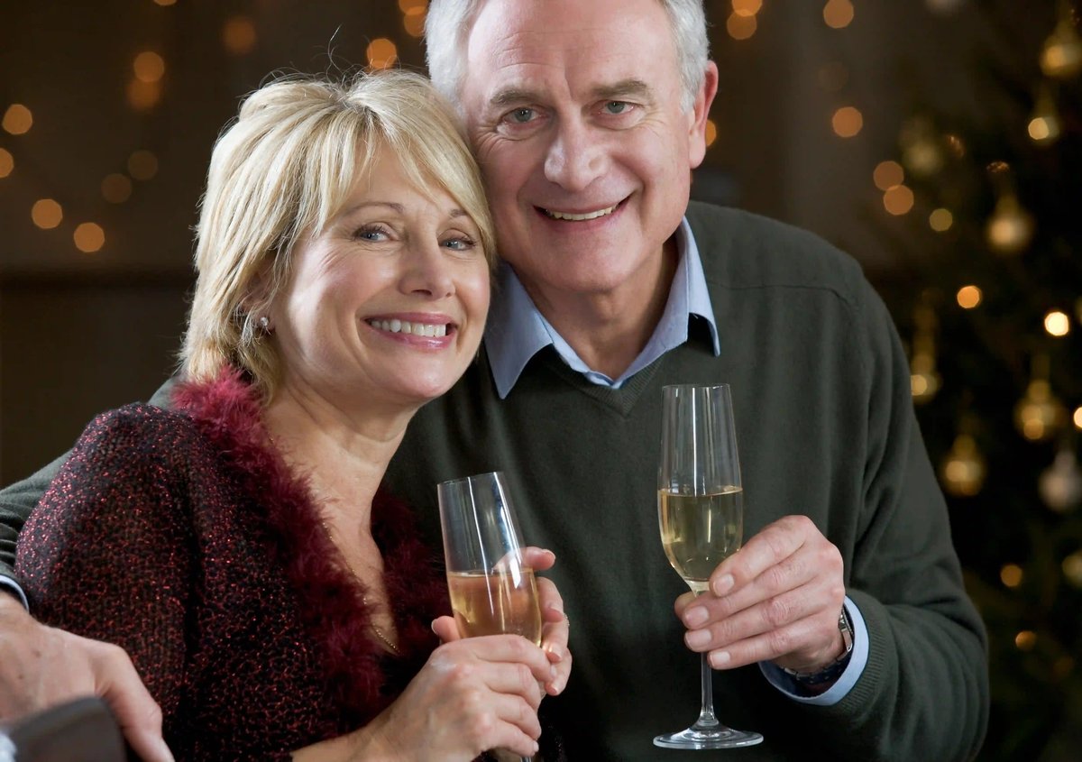Couple souriant trinquant avec des flutes de vin mousseux, lumières chaudes en arrière-plan pour une ambiance de Noël.
