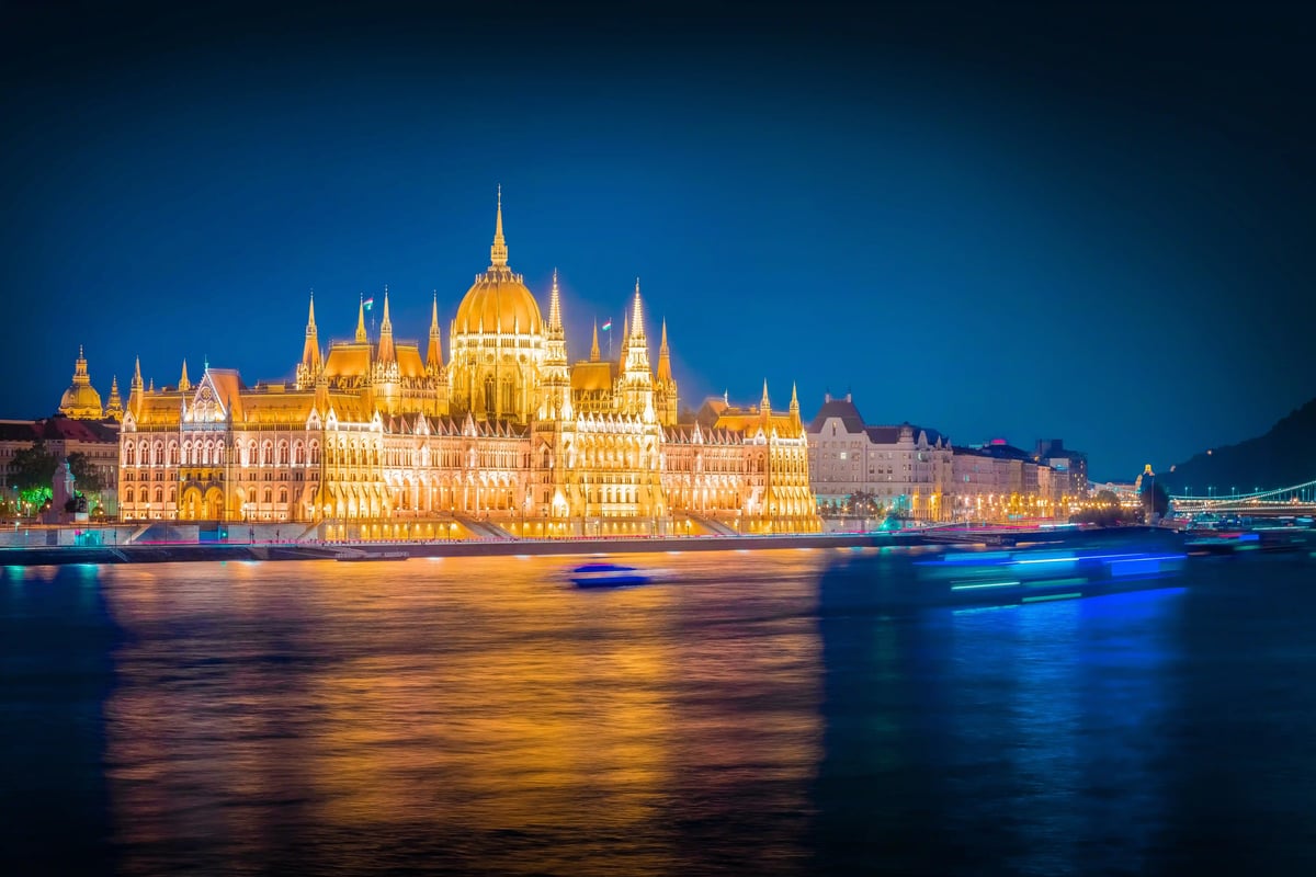 Beleuchtetes Parlamentsgebäude in Budapest bei Nacht am Ufer der Donau mit Spiegelungen im Wasser und vorbeifahrenden Schiffen.