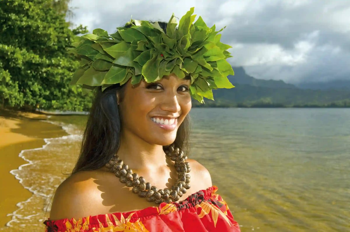 Frau mit traditionellem Blumenschmuck steht lächelnd an einem tropischen Strand